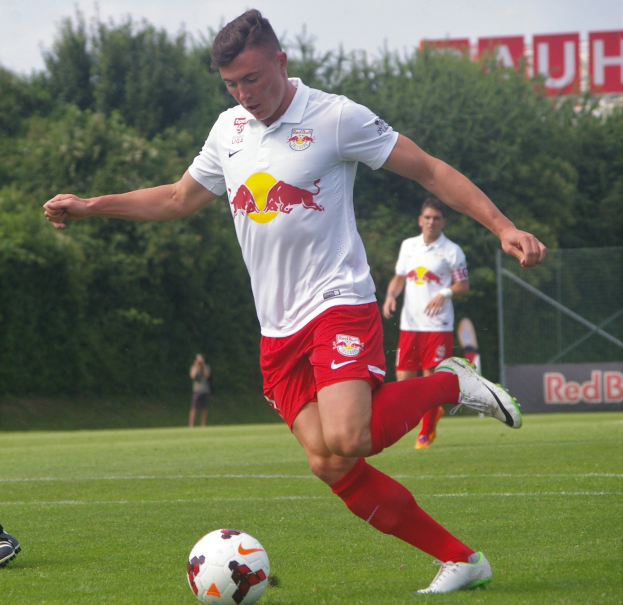 Ein professioneller Füßballspieler schießt einen Ball auf einem grünen Feld mit Bäumen und einem klaren blauen Himmel im Hintergrund, in der Nähe einer Tafel mit der Aufschrift "RB Leipzig."