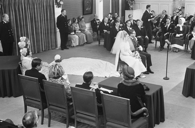 Schwarzes und weißes Foto einer Hochzeitszeremonie mit sitzenden und stehenden Gästen, einem Mann, der in ein Mikrofon spricht, und einer Frau, die einen Strauß hält, mit einem Podium, Blumenschmuck, einem Vorhang, einem Rahmenfoto an der Wand und einer Tür im Hintergrund.