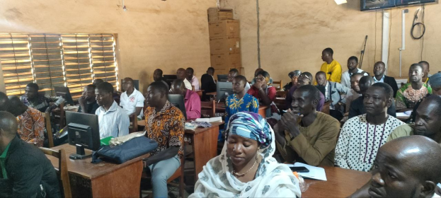 A group of people seated at desks in a classroom with monitors, bags, books, and papers, during a technology training session; a window is on the left, and cardboard boxes and a television are mounted on the wall in the background.