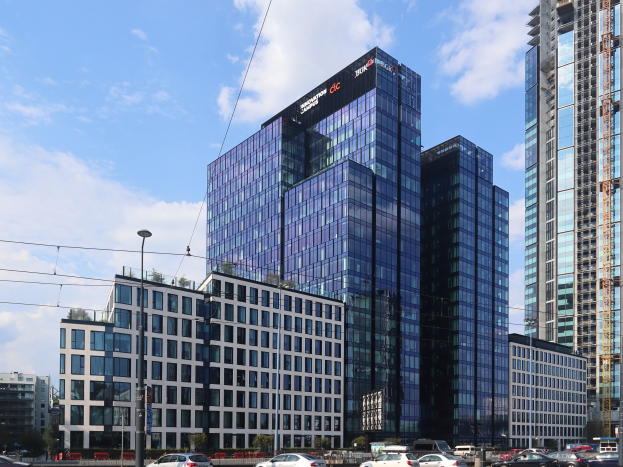 Großes modernes Gebäude mit zahlreichen Fenstern, als Deutsche Bank Hauptsitz in Frankfurt, Deutschland identifiziert, umgeben von städtischer Infrastruktur einschließlich Straßenlaternen, Fahrzeugen, Bäumen und einem bewölkten Himmel.