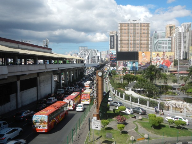 Eine belebte Stadtstraße mit hohen Gebäuden, Fahrzeugen, einer Brücke mit Fußgängern, Bäumen, Schildern und einem bewölkten Himmel.