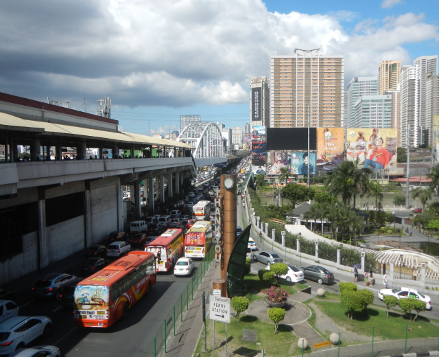 Eine belebte Stadtstraße mit hohen Gebäuden, Fahrzeugen, einer Brücke mit Fußgängern, Bäumen, Schildern und einem bewölkten Himmel.