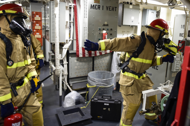 Drei Feuerwehrleute in Schutzausrüstung stehen neben einem Feuerlöscher, mit einer Maschine, einem Müllbehälter, Plastikabdeckungen, Rohren und Lampen im Raum.