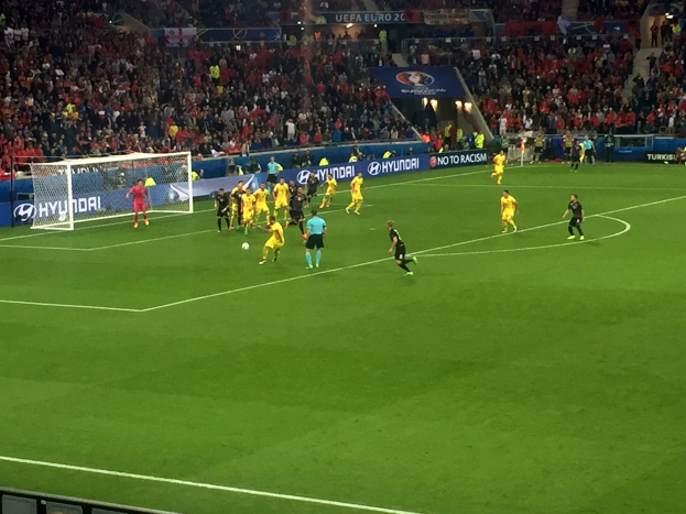 Eine Gruppe von Menschen, die auf einem Feld in einem Stadion Fußball spielen, mit einem Netz auf der linken Seite, einer Menge im Publikum und beleuchteten Tafeln im Hintergrund.