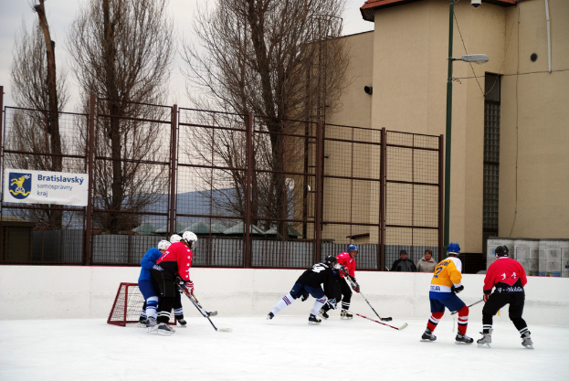 Personen spielen Eishockey auf einer Eisfläche mit Gebäuden, Bäumen, einer Straßenlaterne, einem Namensschild und Zäunen im Hintergrund unter dem Himmel.