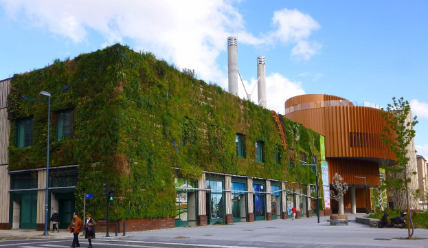Ein große grünwandiges Gebäude in einer urbanen Umgebung mit Fahrzeugen, Füpfen, Straßeninfrastruktur, Bäumen und einem bewölktem Himmel.