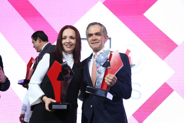 Zwei Menschen mit strahlendem Lächeln und Trophäen auf einer Bühne mit einem Bildschirm im Hintergrund.