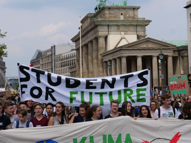 Gruppe von Schülern marschiert in Berlin mit einer bunten "Students for Future"-Schriftzug vor Gebäuden, Bäumen und Himmel.
