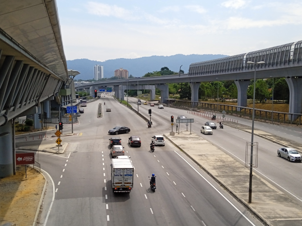 Eine stark befahrene Autobahn mit Schwerlastverkehr neben einer Brücke, mit Straßenlaternen, Ampeln, Verkehrsschildern, Bäumen, Gebäuden, Hügeln und einem bewölkten Himmel.