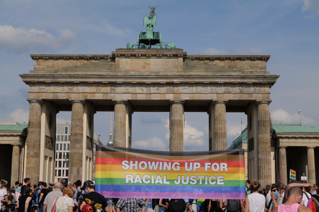 Eine Gruppe von Menschen steht vor dem Brandenburger Tor in Berlin, Deutschland, und hält eine "Racial Justice"-Schleife mit den Säulen und der Statue des Tors im Hintergrund.