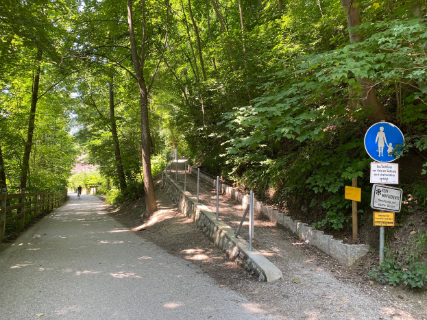 Eine Person fährt mit dem Fahrrad auf einem asphaltierten Weg durch den Wald, umgeben von Bäumen und einem hölzernen Zaun auf der linken Seite und Schildern mit Text und Bildern auf der rechten Seite.