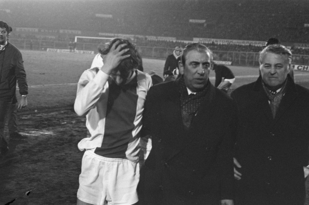 Schwarzes und weißes Bild einer Gruppe von Männern auf einem Fußballfeld mit einem Stadion im Hintergrund, die nach einem gewonnenen Spiel enttäuscht aussehen.