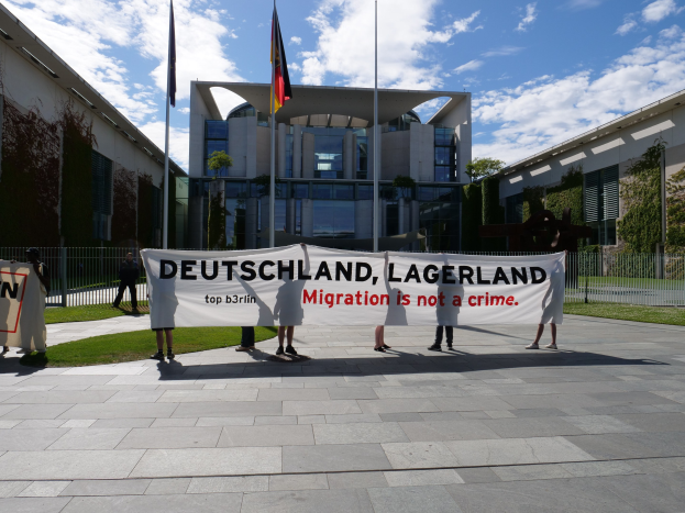 Eine Gruppe von Menschen mit einem Transparent mit der Aufschrift "Deutschland, Lagerland Migration ist kein Verbrechen" vor einem Gebäude mit Fenstern, umgeben von Gras, einem Metallzaun, Pflanzen, Bäumen und Fahnen auf Stangen unter einem bewölkten Himmel.