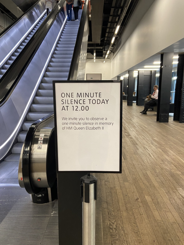 Eine Rolltreppe in einem Flughafen mit einem Schild, auf dem "Eine Minute Stille heute" steht, ein paar Menschen darauf und Lichter an der Decke im Hintergrund.