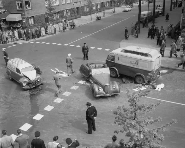 Schwarz-weißes Bild einer Autounfall-Szene an einer Straßenkreuzung mit mehreren Fahrzeugen, darunter ein Van, und einer Gruppe von Menschen, die daneben stehen, mit Gebäuden, Bäumen, Pfählen und Texttafeln im Hintergrund.