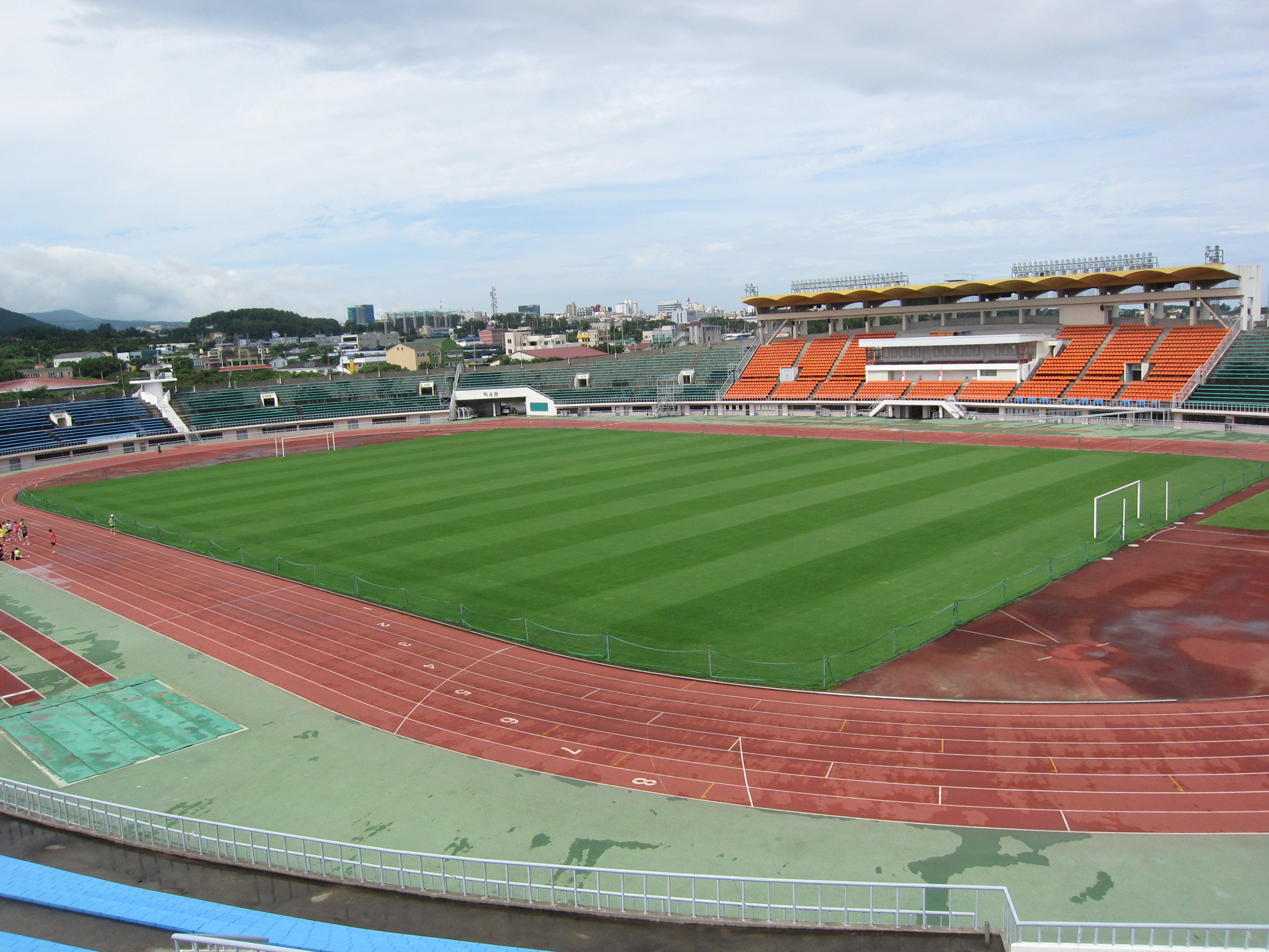 Großes Stadion mit einem Fußballfeld, umgeben von Gebäuden, Bäumen, Hügeln und einem klaren blauen Himmel, mit ein paar Menschen auf saftig grünem Rasen.