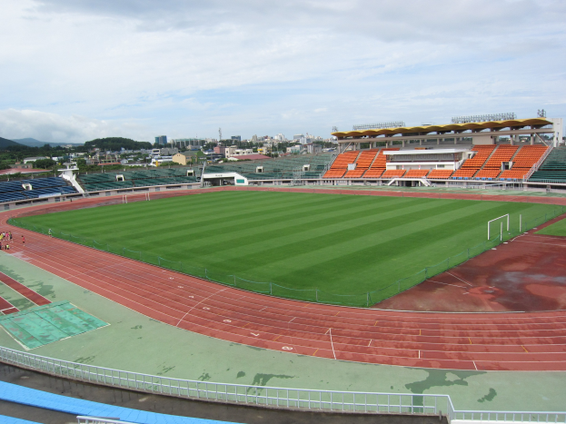 Großes Stadion mit einem Fußballfeld, umgeben von Gebäuden, Bäumen, Hügeln und einem klaren blauen Himmel, mit ein paar Menschen auf saftig grünem Rasen.