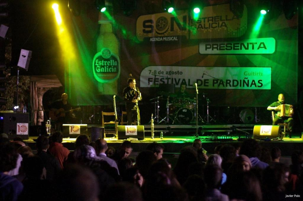 Publikum schaut auf der Bühne mit Musikinstrumenten, Lautsprechern, Mikrofonen, einer Kulisse und Scheinwerfern, mit einer Flasche und anderen Gegenständen im Hintergrund.