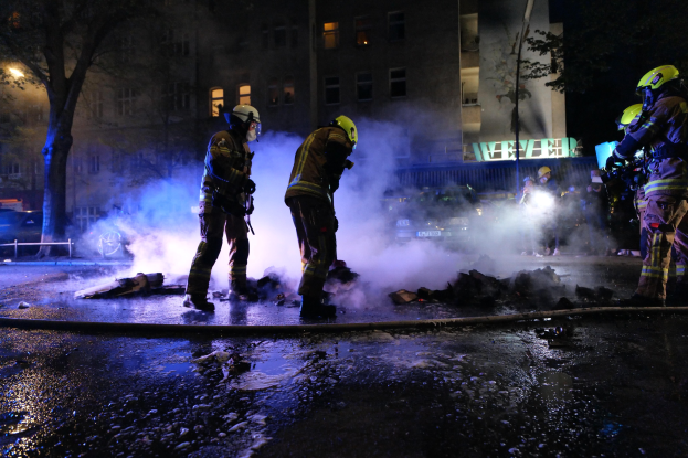 Feuerwehrleute in Helmen stehen nachts auf einer Straße mit Rauch aus einem Rohr aufsteigend, haltend Gegenstände, während Gebäude, Bäume und Fahrzeuge im Hintergrund mit einer Tafel auf der rechten Seite sichtbar sind.