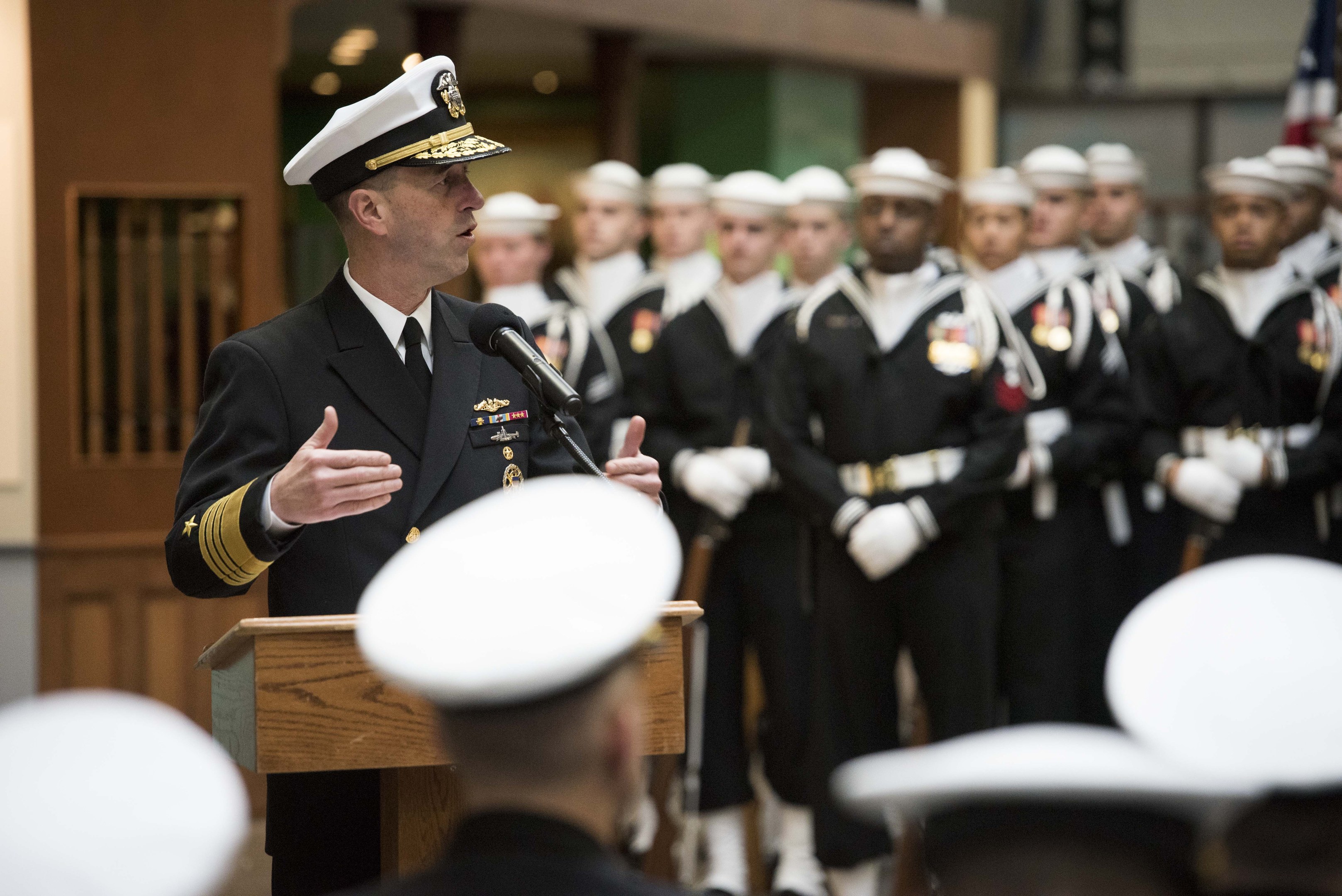 Ein Mann in Uniform spricht vor einem Podium während einer Abschlussfeier, während uniformierte Personen im Hintergrund Gewehre halten.