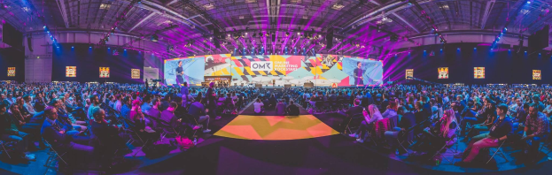 Eine große Zuschauermenge sitzt auf Stühlen einem hell erleuchteten Podium mit mehreren Lautsprechern und Bildschirmen gegenüber, wahrscheinlich bei einem Online-Marketing-Event.