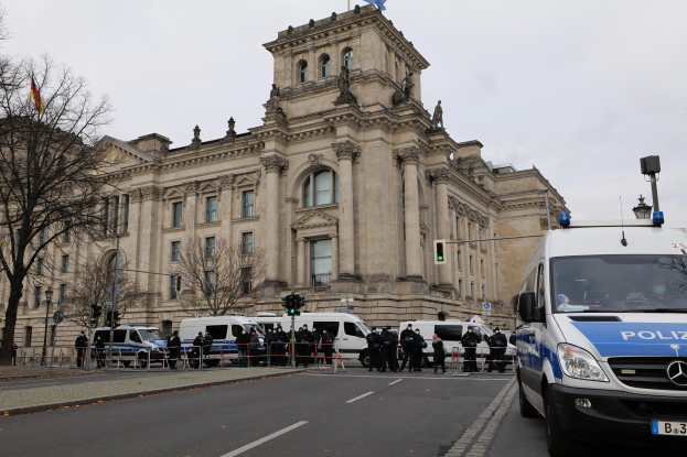 Eine Gruppe von Polizisten vor dem Reichstaggebäude in Berlin, Deutschland, mit Fahrzeugen, einer Absperrung, Verkehrsampeln, Laternen, Bäumen und Flaggen im Hintergrund.