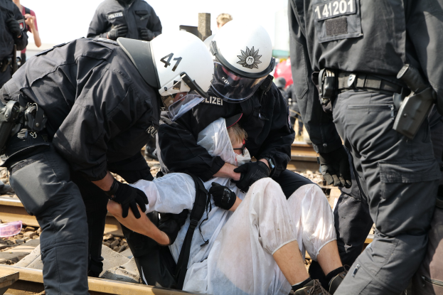 Eine Gruppe von Polizisten in Einsatzkleidung nimmt einen Mann in einem weißen Kleid auf einem Bahngleis fest, mit anderen im Hintergrund.