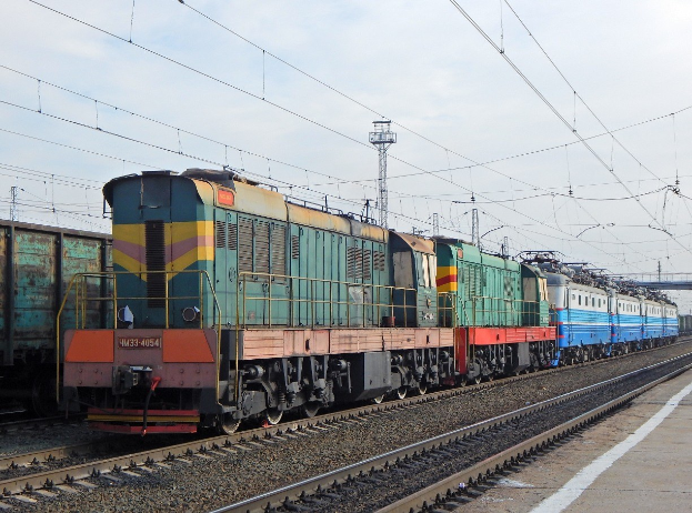 Ein diesel-elektrisches Lokomotiv fährt auf Schienen neben einer Plattform, mit elektrischen Polen und Drähten im Hintergrund unter einem klaren Himmel.