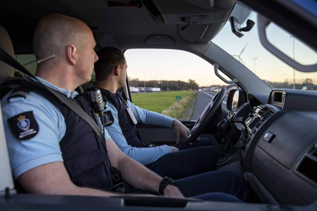 Zwei Polizeibeamte sitzen in einem Auto auf einer Straße mit Gras, Bäumen und Polen, unter einem klaren blauen Himmel.