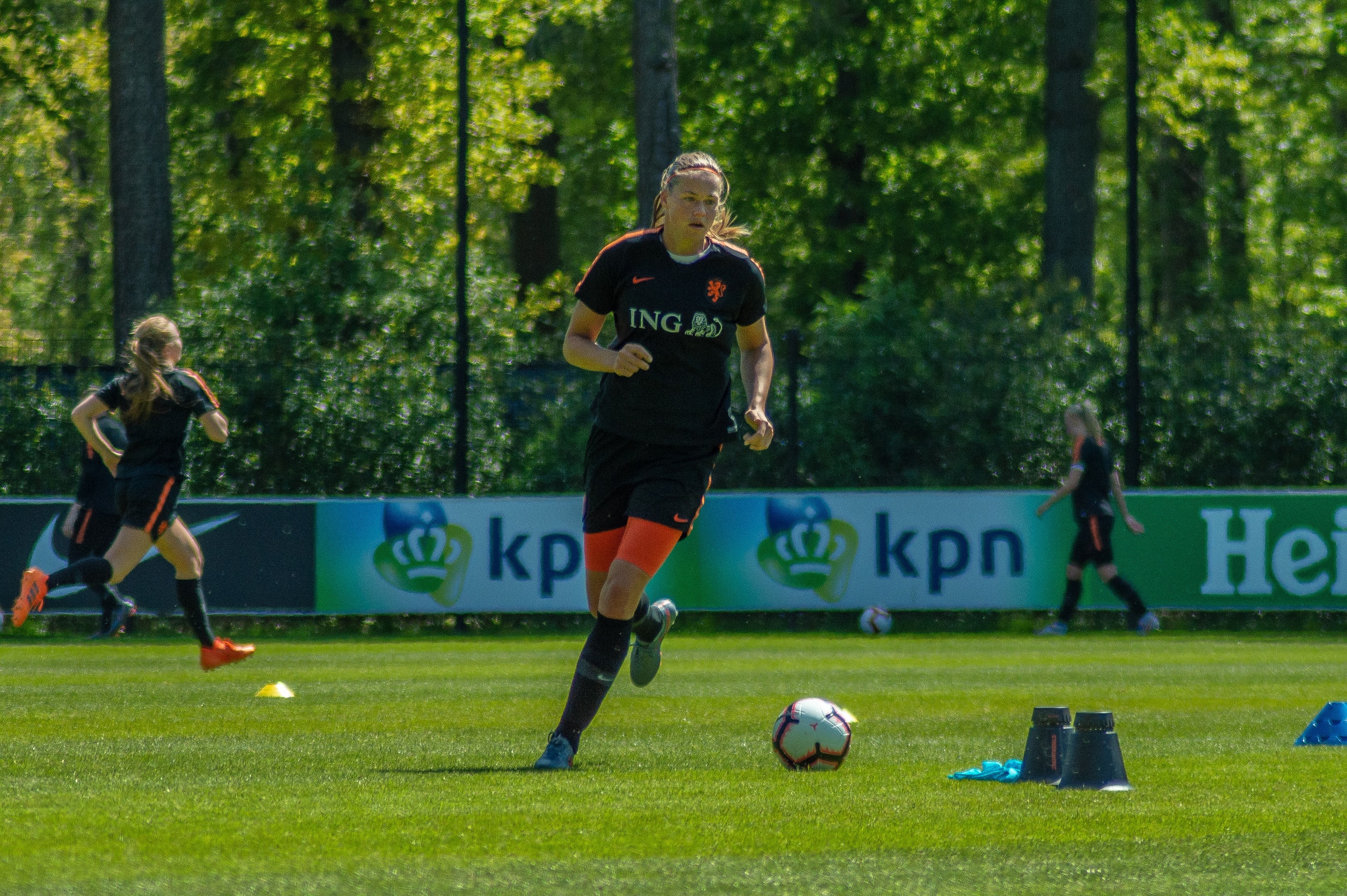 Gruppe von Frauen in Fußballtrikots beim Fußballspielen auf einem grünen Feld mit Bäumen und Pfosten im Hintergrund, ein Banner ist im Hintergrund sichtbar.