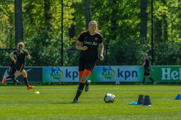 Gruppe von Frauen in Fußballtrikots beim Fußballspielen auf einem grünen Feld mit Bäumen und Pfosten im Hintergrund, ein Banner ist im Hintergrund sichtbar.