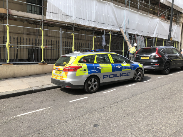 Polizeiauto auf einer Straße mit zwei Helmträgern und einem weiß verhüllten Gebäude im Hintergrund.