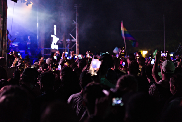 Eine Menge bei einem Konzert hält leuchtende Handys hoch, während Musiker auf der Bühne unter hellen Lichtern spielen, umgeben von Pfählen und einer Fahne.