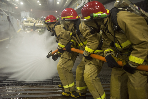 Feuerwehrleute in Helmen und Handschuhen sprühen Wasser auf ein Feuerwehrauto, während sie in der Nähe von Equipment und Beschilderung stehen.