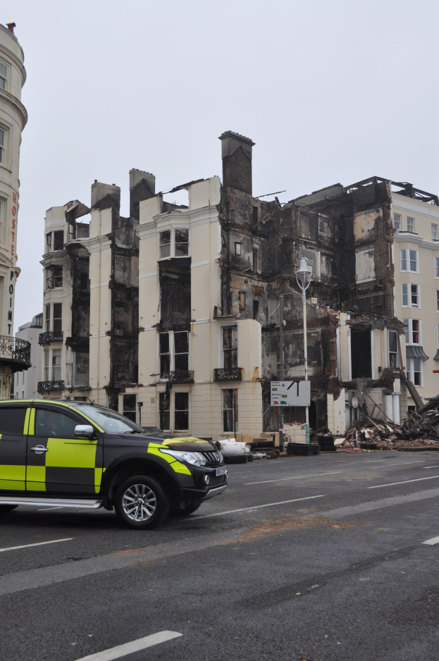 Ein Polizeiauto vor einem schwer beschädigten Gebäude mit zersplitterten Fenstern und verstreuter Trümmer, unter einem sichtbaren Himmel mit nahen Gebäuden.