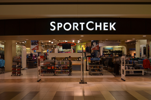 Außen- und Innenansicht eines Sportchek-Verkaufsstellen in einem Einkaufszentrum, mit Kleiderstangen, Deckenleuchten, Säulen und einem Fernseher.