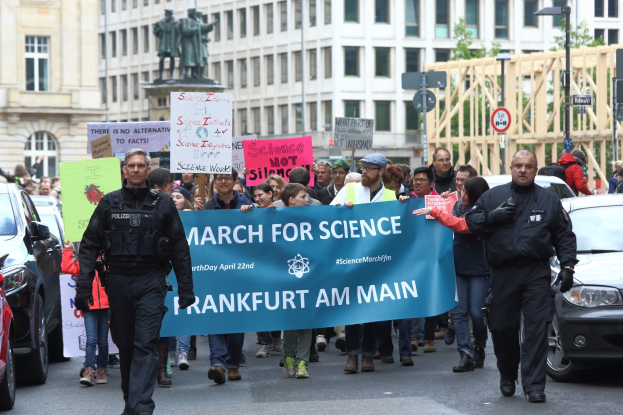 Gruppe von Menschen marschiert mit einem "March for Science Frankfurt am Main"-Schild die Straße entlang, während Autos neben ihnen fahren und Gebäude, Statuen, Laternenpfähle, Schilder und Bäume im Hintergrund zu sehen sind.