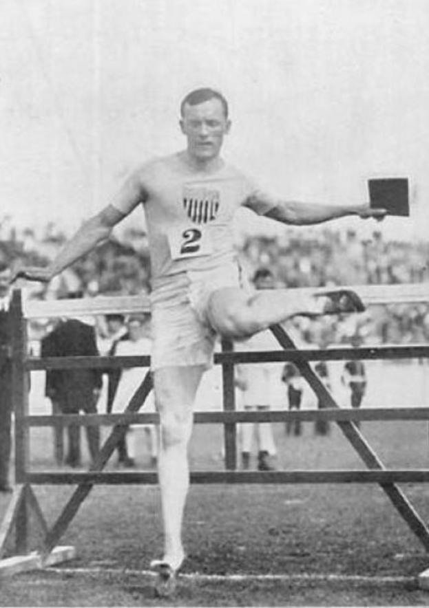 Schwarz-weißes Foto eines Mannes in der Luft, der über ein Hindernis springt, mit einer nummerierten Weste und Shorts, umgeben von Zuschauern und Himmel.