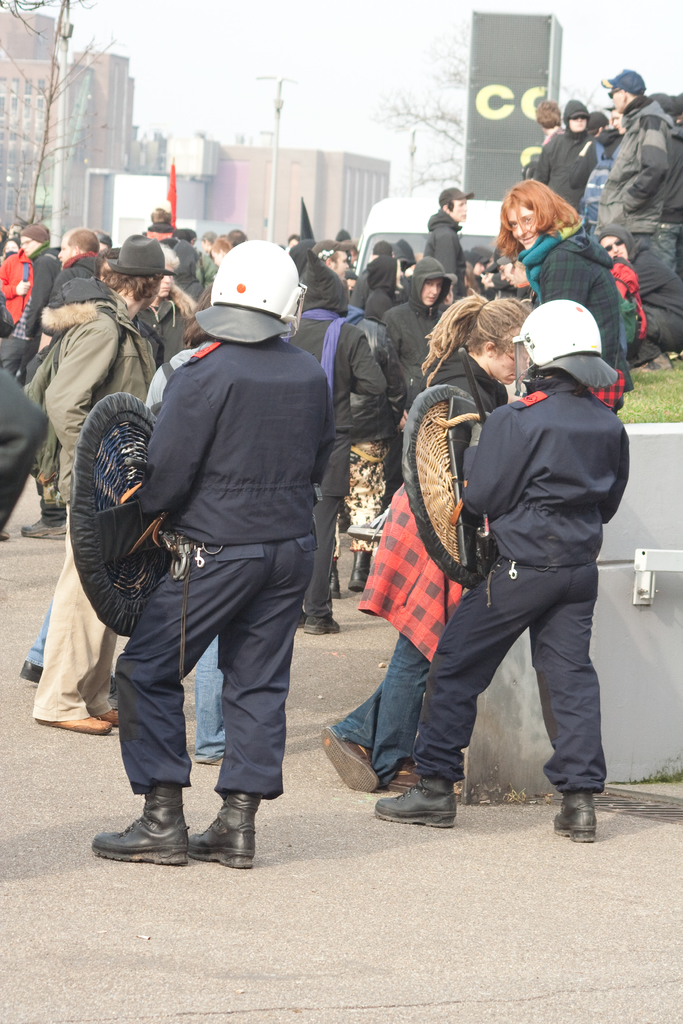 Eine Gruppe von Menschen, die auf einer Straße gehen, mit zwei Personen in der Vordere, die wie Polizisten aussehen, Gebäude im Hintergrund und Boden unten.