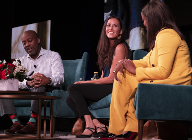 Drei Personen sitzen auf einem Sofa vor einem Bildschirm, mit einer Vase und einer Flasche auf dem Tisch dazwischen, während einer Podiumsdiskussion über Geschlechtergerechtigkeit und Frauenrechte.