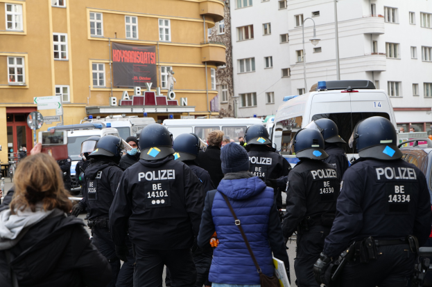 Eine Gruppe von Polizisten vor einer Menge mit Helmen und Jacken während einer Demonstration in Berlin, mit Fahrzeugen, Gebäuden, Laternenmasten und einem Banner im Hintergrund und einer Person mit einer Kamera.