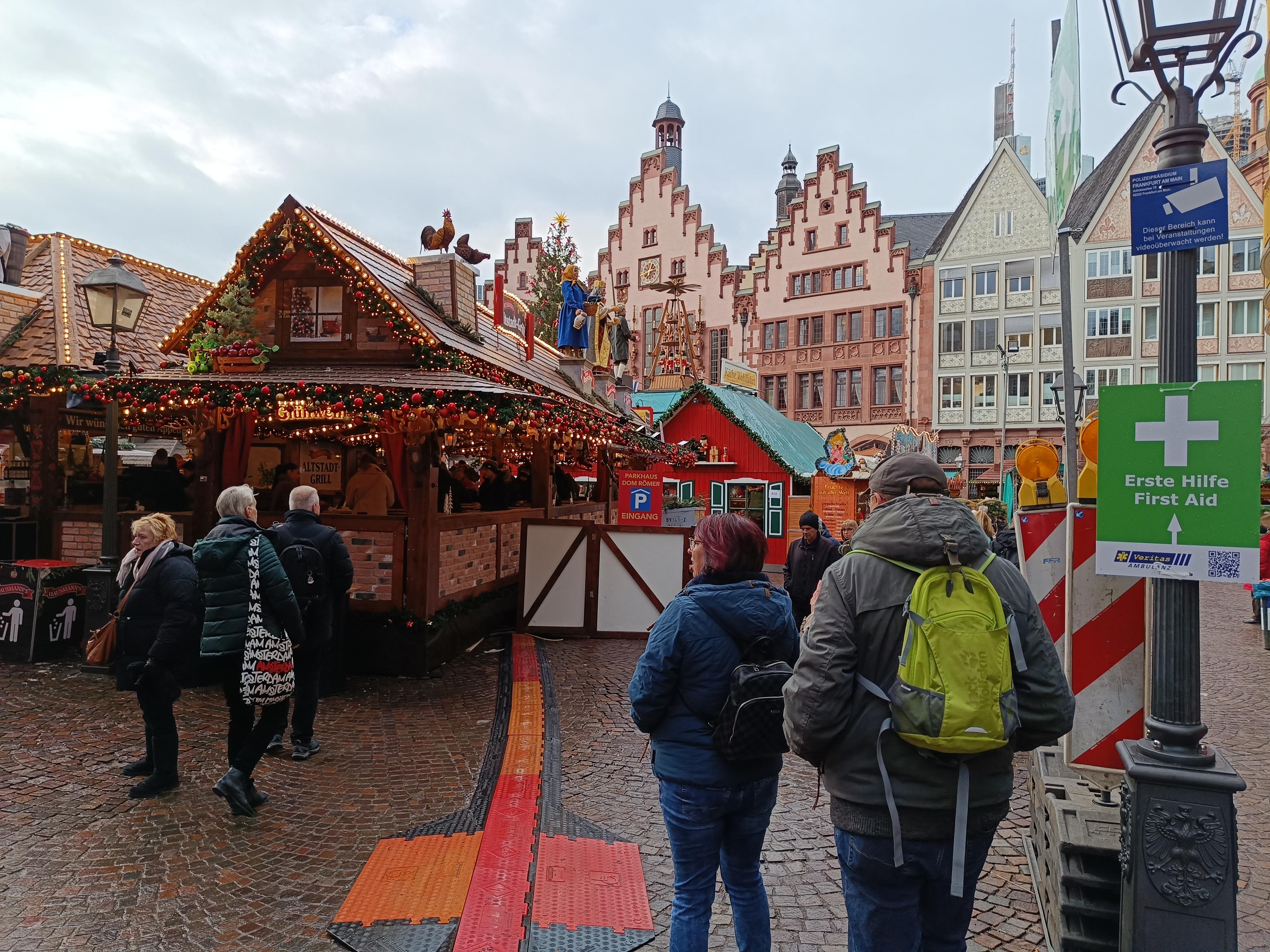 Eine Gruppe von Menschen geht eine Kopfsteinpflasterstraße neben einem Weihnachtsmarkt in Nürnberg, Deutschland, entlang, einige tragen Taschen, Laternenmasten und Texttafeln, während Gebäude mit Fenstern und ein bewölkter Himmel im Hintergrund zu sehen sind.