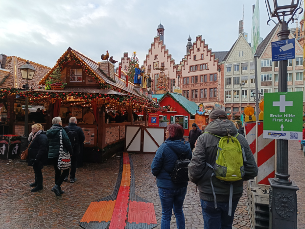Eine Gruppe von Menschen geht eine Kopfsteinpflasterstraße neben einem Weihnachtsmarkt in Nürnberg, Deutschland, entlang, einige tragen Taschen, Laternenmasten und Texttafeln, während Gebäude mit Fenstern und ein bewölkter Himmel im Hintergrund zu sehen sind.