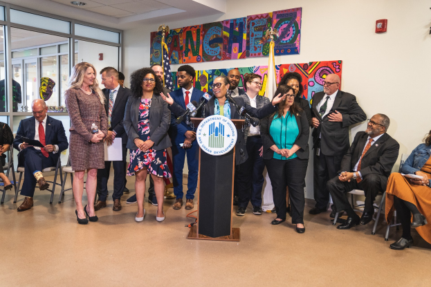 Gruppe von Menschen vor einem Rednerpult mit Mikrofon und einer Tafel mit der Aufschrift "Sancho-Stadtrat tritt dem Stadtrat bei", mit sitzenden und stehenden Teilnehmern, Fahnen, Feuerlöscher und Deckenbeleuchtung.