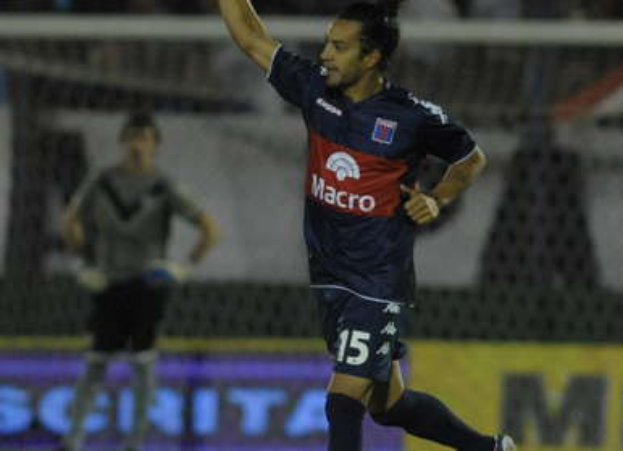 Ein Fußballspieler feiert nach einem Tor auf dem Boden vor einer jubelnden Menge und einer Person im Hintergrund sowie einer Tafel mit Text dahinter, alles mit einem unscharfen, dynamischen Hintergrund.