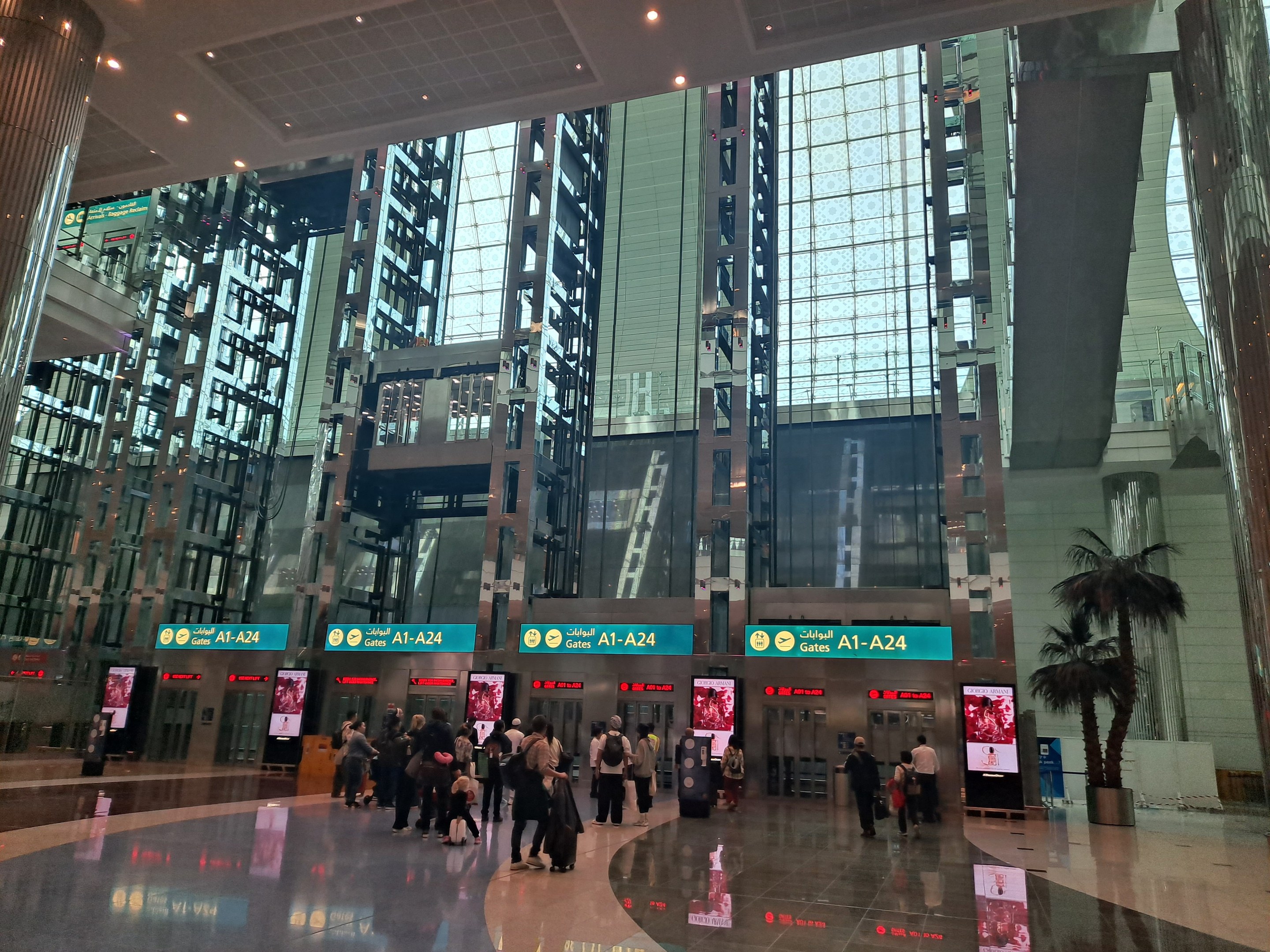 Menschen mit Gepäck durch eine Flughafenhalle gehen, umgeben von Gebäuden, Bäumen, Texttafeln, Säulen und Deckenleuchten.