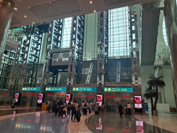 Menschen mit Gepäck durch eine Flughafenhalle gehen, umgeben von Gebäuden, Bäumen, Texttafeln, Säulen und Deckenleuchten.