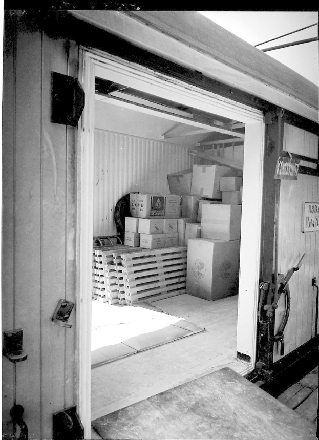 Schwarze und weiße Fotografie eines metallenen Seecontainers mit einer Tür auf der linken Seite, gefüllt mit ordentlich gestapelten Kartons.