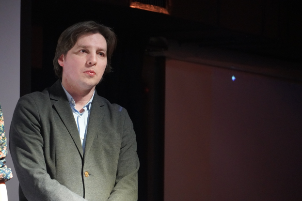 Zwei Personen nebeneinander auf einer Bühne, eine im Anzug und die andere neben ihnen, beide mit ernsten Gesichtern.