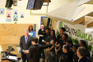 Eine Gruppe von Menschen steht um ein Rednerpult vor einer Menge, mit einem Tisch mit Mikros und Papieren links, Plakaten an der Wand im Hintergrund, Gegenständen auf dem Dach und einem Gegenstand rechts, während sie Hände schütteln, was darauf hinweist, dass sie Teil einer Jugendgruppe sind.